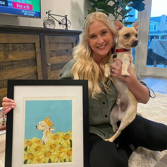 a blonde woman holding a blonde shorthaired chihuahua and posing with a framed painting of a chihuahua wearing a crown in a field of flowers
